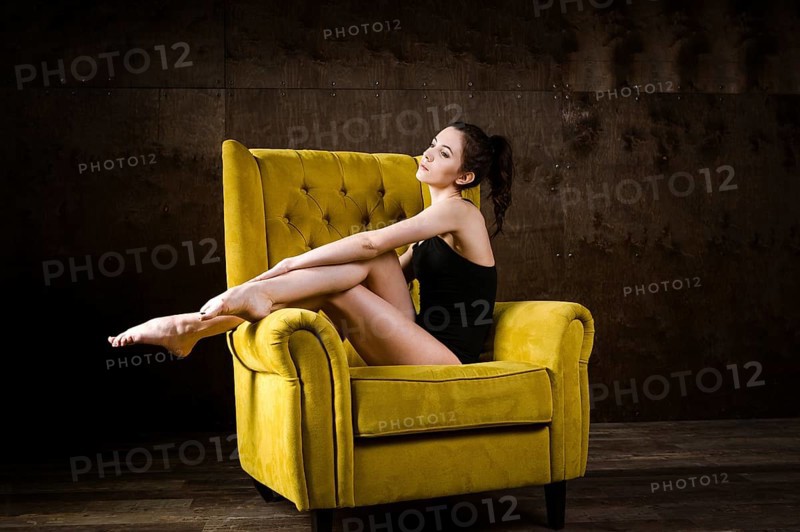 A young beautiful, sexy Caucasian woman with thin figure and long bare legs, barefoot posing reclining on yellow armchair in the interior against dark wooden wall. Dressed in a black classic swimsuit