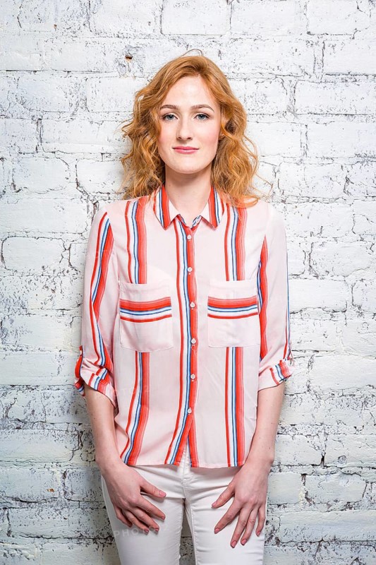 A beautiful young woman student with red curly hair and freckles is leaning against a brick wall of grey colour. Dressed in a red striped shirt and white pants