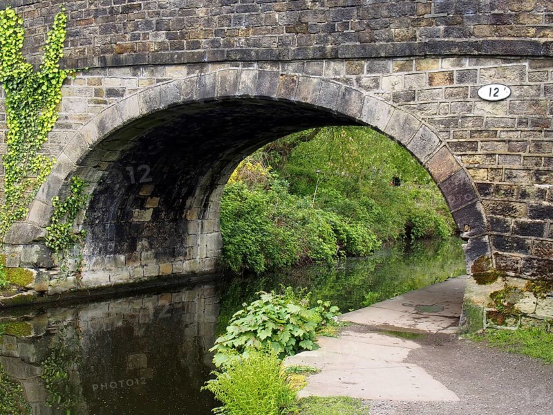 An old stone bridge crossing the rochdale canal at mytholmroyd west yorkshire with a waterside pathway surrounded by trees and bushes
