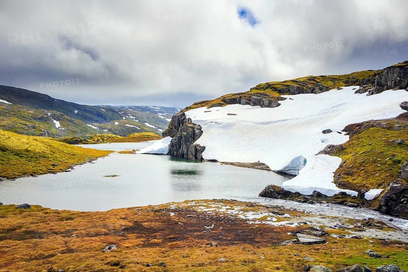The cold water of the lake is covered with ice floes. Gorgeous Snow Road in Norway. Cold July in Northern Europe. The cold snowfields and sharp stone placers