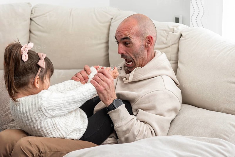 Bald father playing with his daughter on the sofa, making funny faces and holding her feet
