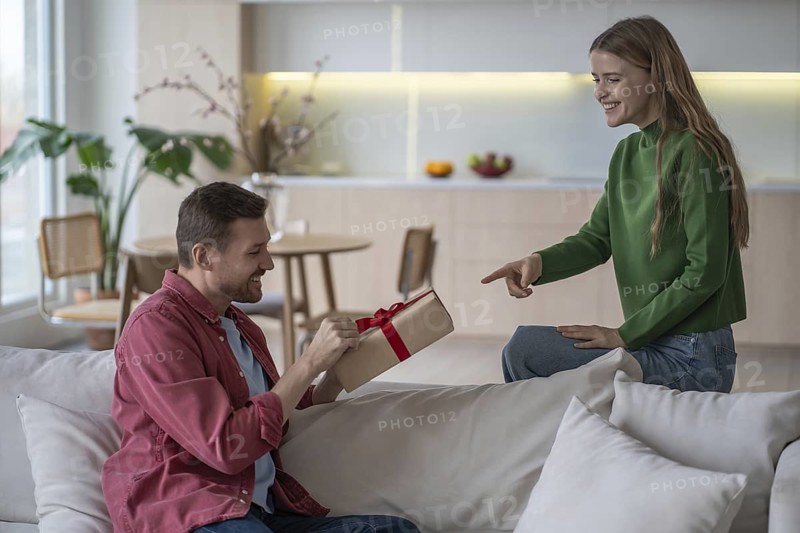 Happy couple celebration. Loving pleased woman gives gift with red tape to smiling husband. Satisfied man holding present box from girlfriend on birthday feeling interest and anticipation at home
