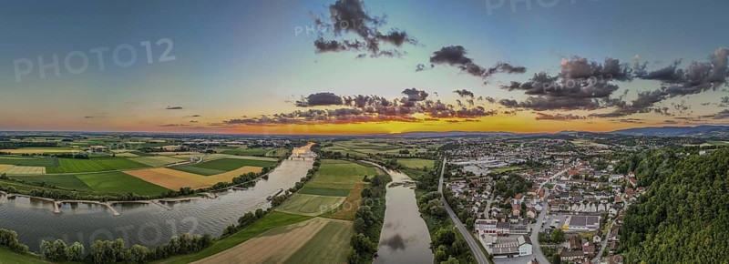 The sunset bathes the landscape in warm light, while the Danube flows calmly and the town of Bogen (Lower Bavaria) is visible in the background. Green fields and rolling hills complete the panorama
