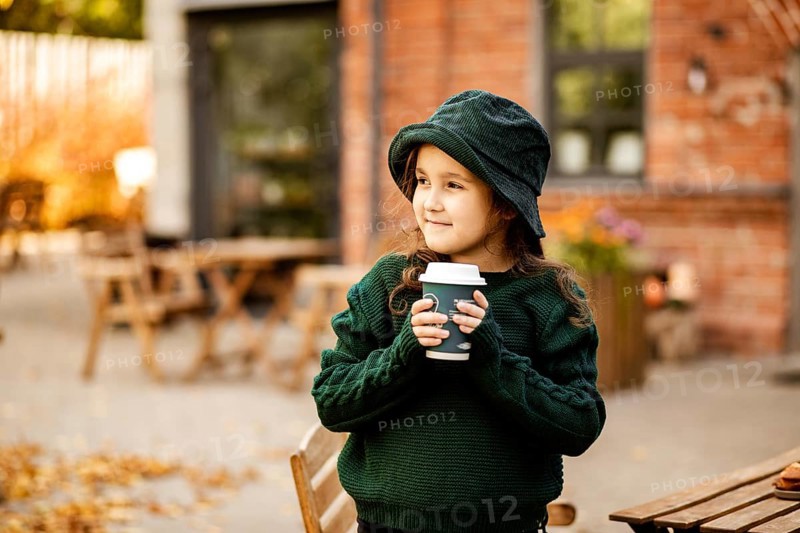 Cute little girl drinking hot cocoa from paper cup in outdoor cafe or backyard. Autumn concept