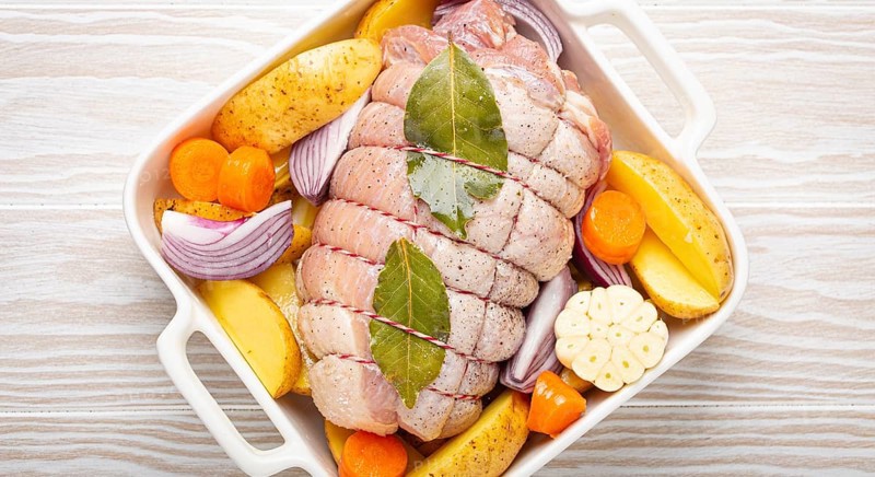 Raw wrapped rolled sliced pork in white casserole dish with potatoes, vegetables and herbs on rustic white wooden background top view. Pork roll with vegetables ready to be prepared, food photography, food photography