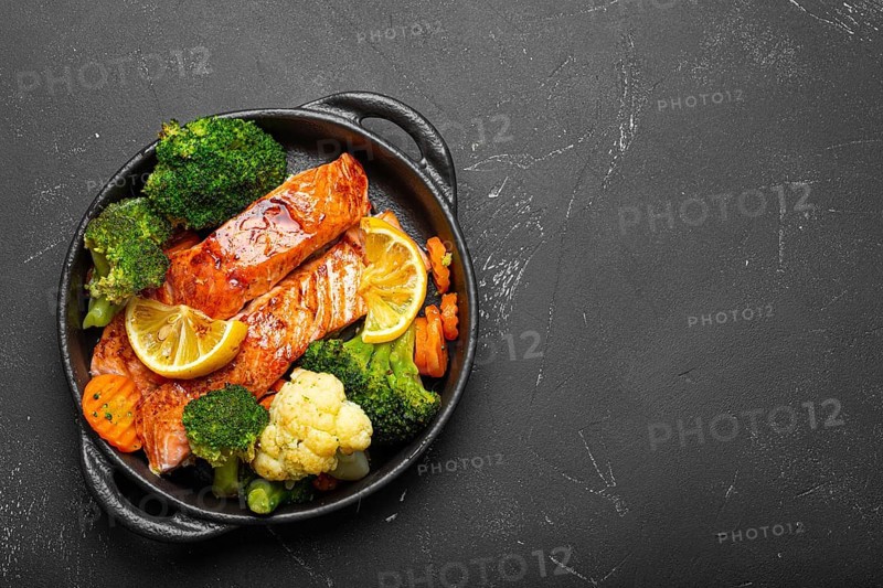 Healthy baked fish salmon steaks, broccoli, cauliflower, carrot in cast iron casserole bowl black dark stone background. Cooking a delicious low carb dinner, healthy nutrition, food photography, food photography