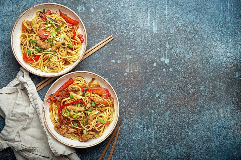 Two bowls with Chow Mein or Lo Mein, traditional Chinese stir fry noodles with meat and vegetables, served with chopsticks top view on rustic blue concrete background, space for text, food photography, food photography