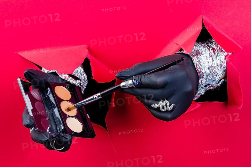 Hands with black gloves holding brush and powder box. Breaking through a red paper wall