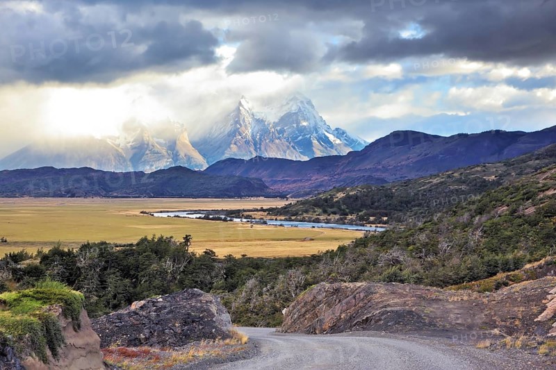 The road between turned yellow hills and river goes to snow-covered black rocks. Patagonia. Vertiginous landscape in the Chilean Andes