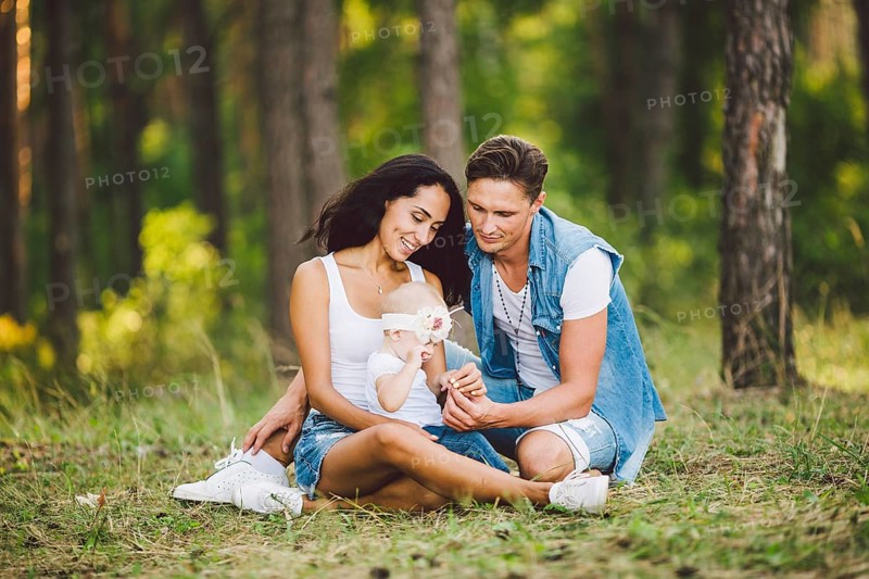 The theme family outdoor recreation in forest park area. Young beautiful stylish caucasian family with small child daughter one year old sit on the graß in the coniferous forest in summer at sunset
