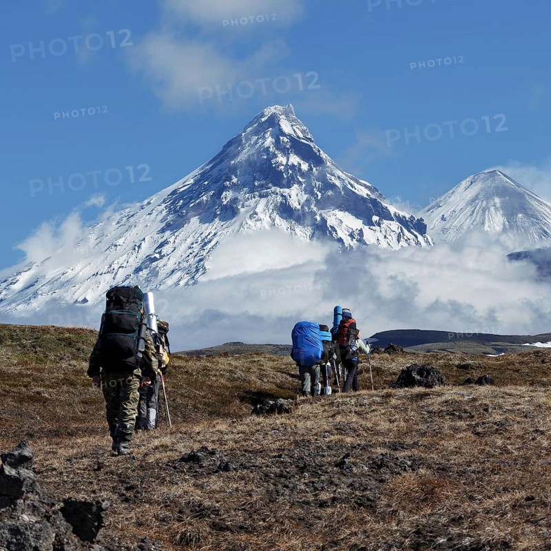 Hiking on Kamchatka Peninsula: group of tourists and travelers with backpacks behind the shoulders climbing to mountains on background of rocky Klyuchevskaya group of volcanoes on sunny day. Kamchatka Peninsula, Russian Far East