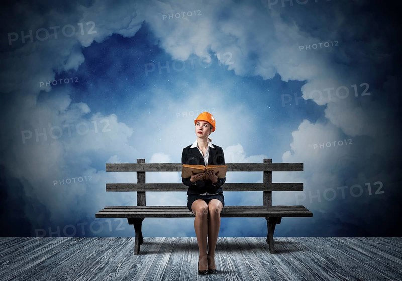 Beautiful woman architect sitting on wooden bench. Young technician specialist in orange safety helmet holding open book in hands on cloudy night sky background. Engineering and development company