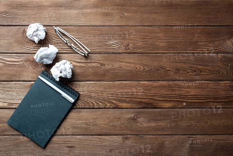 Top view of well organized home office workplace. Black spiral notepad, crumpled paper and glasses on brown wooden surface. Education, creativity and working minimalist concept with copy space