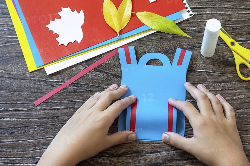 Instruction step 6. Greeting card paper backpack on a wooden table. Handmade. Project of children's creativity, handicrafts, crafts for kids