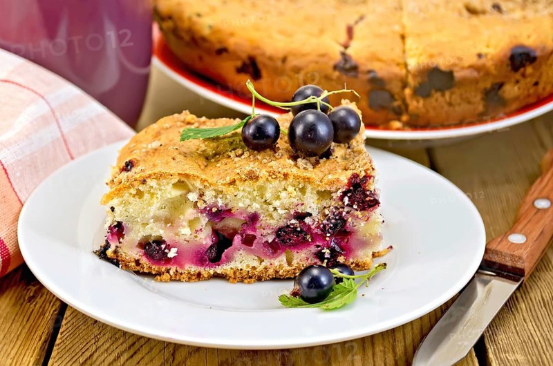 Sweet piece of cake with black currant, napkin, cup, knife on background wooden plank