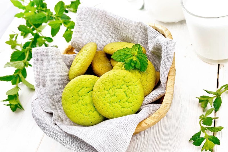 Green mint cookies on napkin in wicker basket, milk in a glass and a jug on white wooden board background