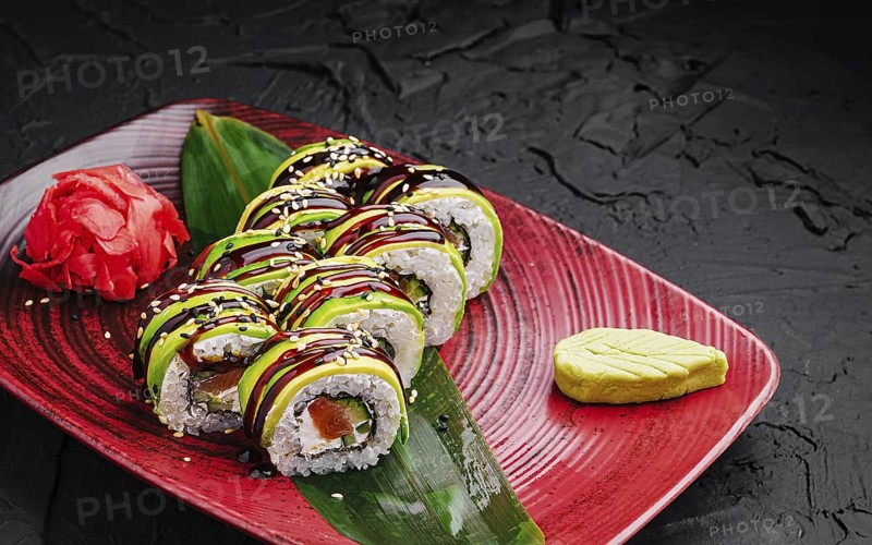 Food, Delicious sushi rolls are beautifully arranged on a red plate. They feature fresh ingredients, topped with avocado and drizzled with sauce, accompanied by wasabi and pickled ginger