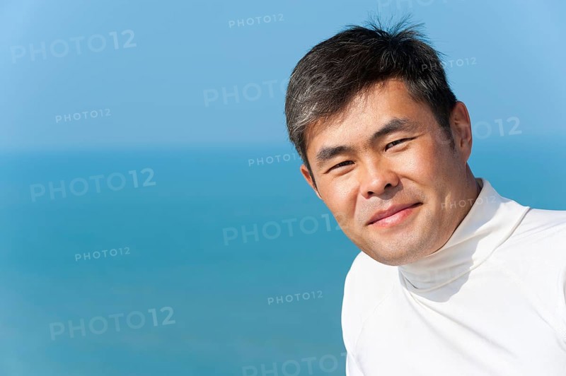 Handsome asian young man looks into camera smiling, blue sea and sky