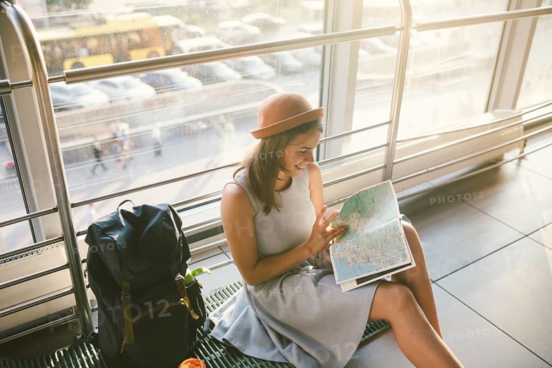Waiting, delayed transport in the terminal of the airport or train station. Young caucasian woman in dress and hat sits on tourist rug with backpack near window to room and looks into tourist map