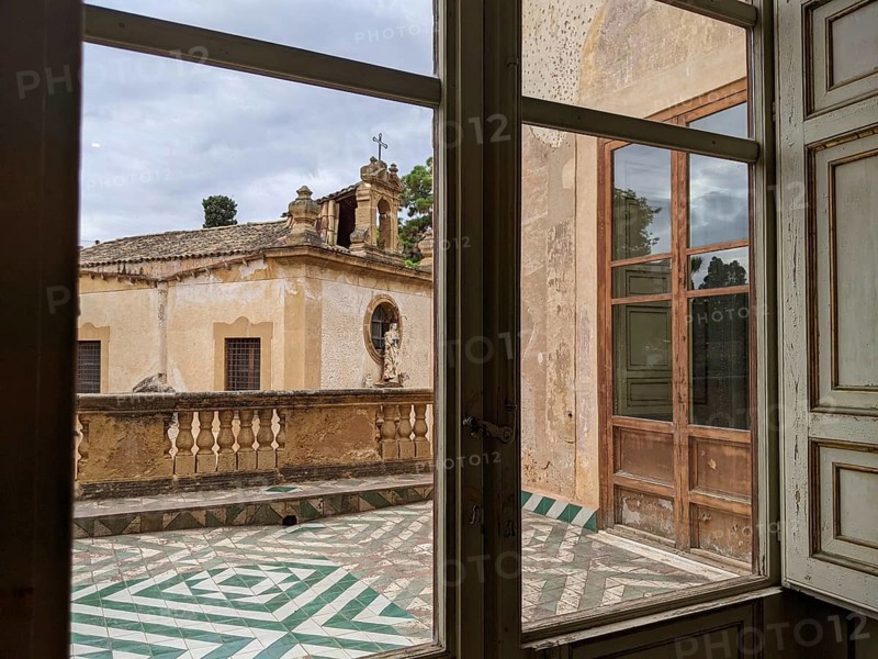 A window view of a historic terrace with patterned floor tiles and vintage architectural elements, Villa Palagonia, known as the ‘villa of monsters', is an 18th-century walled building located in Bagheria, Sicily