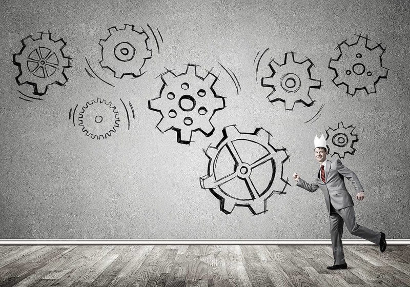 Young handsome businessman wearing white paper crown running and gear mechanism on wall