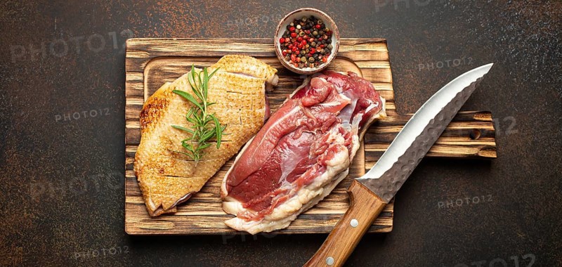 Two raw uncooked duck breast fillets with skin, seasoned with salt, pepper, rosemary top view on wooden cutting board with knife, dark brown concrete rustic background, food photography, food photography
