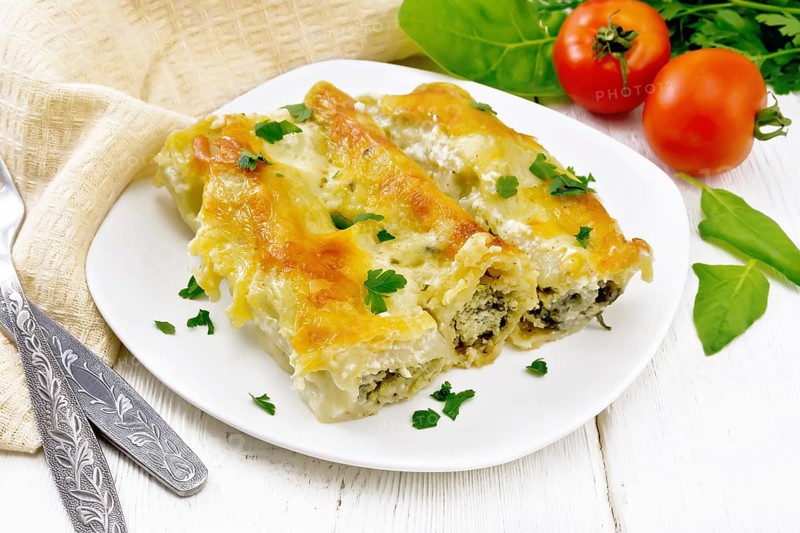 Cannelloni stuffed with cottage cheese and spinach with Bechamel sauce in a plate, towel, tomatoes and parsley on the background of light wooden board