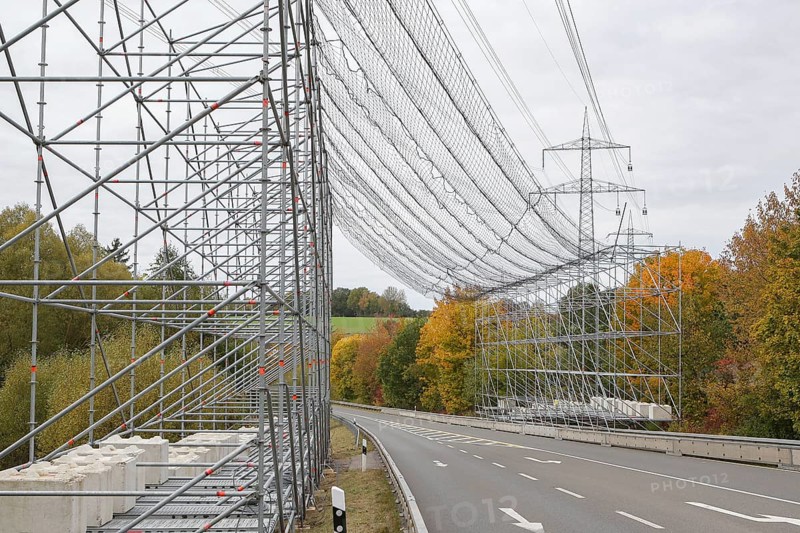 A scaffold with steel net as protection over a road, safety measure due to work on an overhead line and electricity pylons, in the background trees in autumn