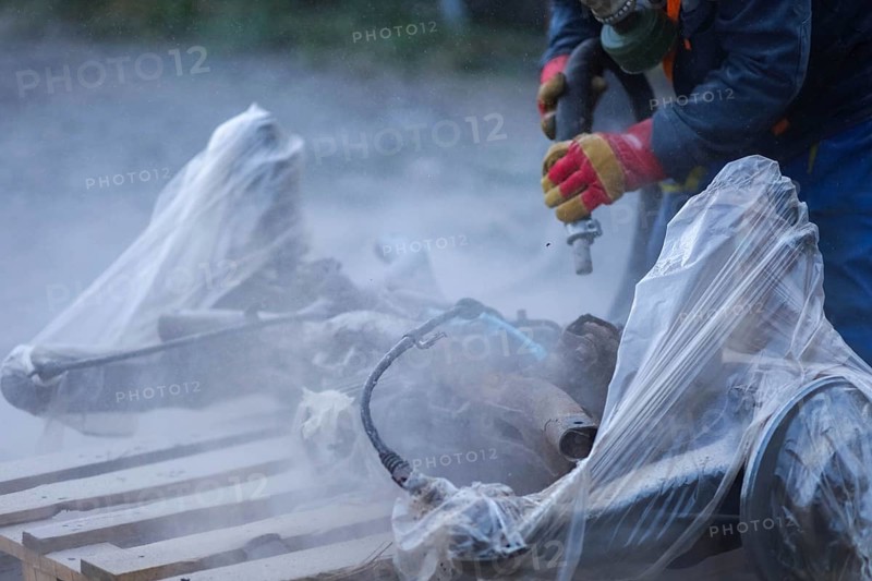 A worker in protective gloves sanding car parts covered in plastic, producing dust