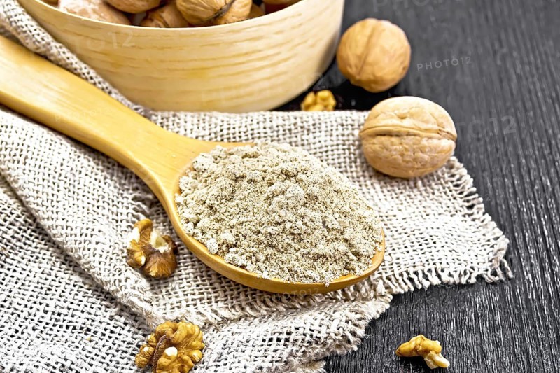 Walnut flour in a spoon on burlap, nuts on a table and in a bowl against dark wooden board