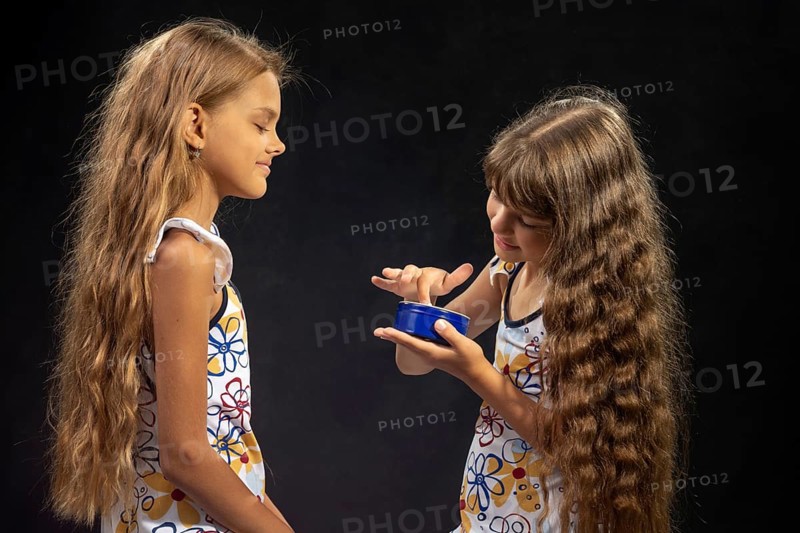 A girl puts jar cream on her finger, another girl sits nearby