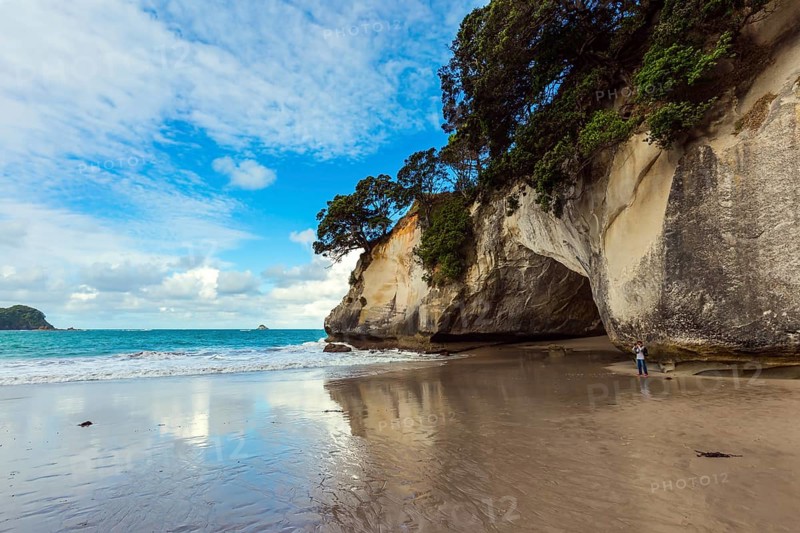 Woman takes pictures of magnificent landscape. Ocean tide in the Cathedral Cove. Mirror reflections of clouds in wet sand. Travel to New Zealand. The concept of exotic, ecological and photo tourism
