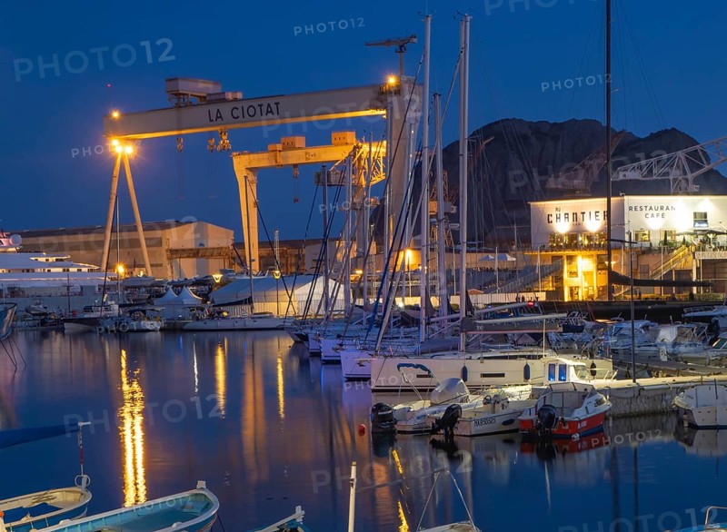 The famous harbour of La Ciotat, France, with fishing and sailing boats, in front of the industrial area with the illuminated dockside crane. Situation in the evening during blue hour
