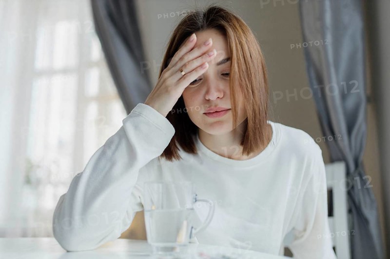 Woman holding her forehead looking fatigued, sitting indoors with a glass of water, suggesting a headache or stress