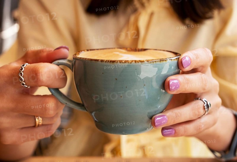 Woman in yellow clothes holding blue cup of cappuccino with both hands, close-up