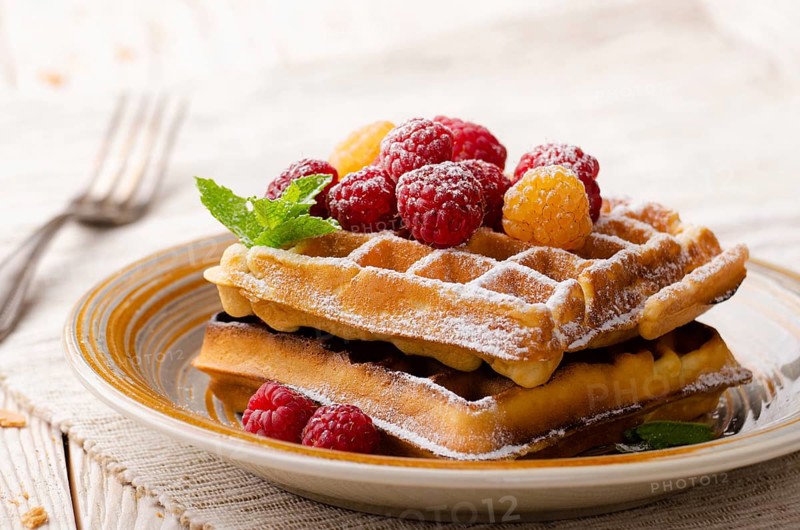 Belgian waffles served with raspberries and mint leaf dusted with powdered sugar on white wooden kitchen table