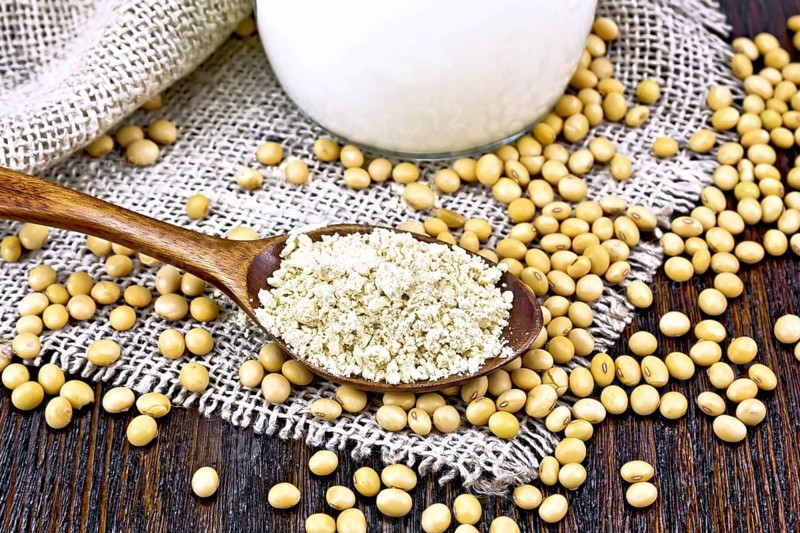 Soy flour in a spoon and milk in glass jug, soybeans on sackcloth on wooden board background