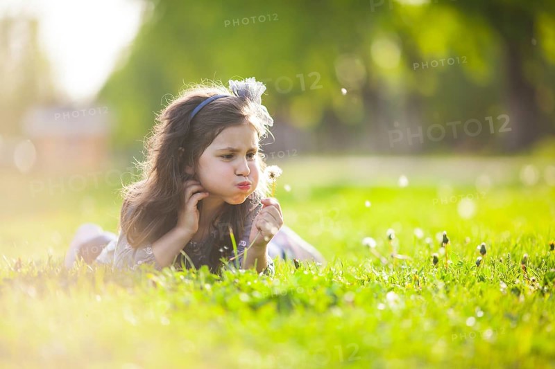 The little girl lays on a grass and blowing dandelion on the summer day