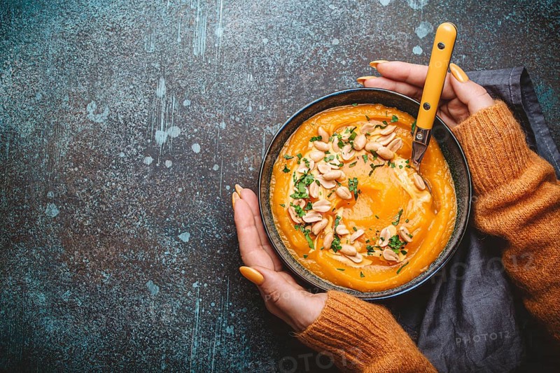 Female hands in yellow knitted sweater holding a bowl with pumpkin cream soup on dark stone background with spoon decorated with peanuts, top view. Autumn cozy dinner concept, space for text, food photography