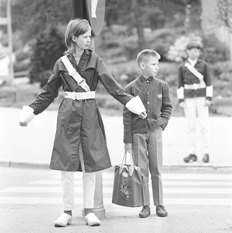 Junior safety patrol in the 1960s. A teenage girl stands with her arms out and signals to the pedestrians to stop at the crosswalk. When the cars have passed or stopped, the children can walk across the street. Being part of the Junior safety patrol was voluntary for the school students and the reward was sometimes to go on shorter trips and excursions. Nowadays, the operation is closed in many municipalities. Sweden 1964