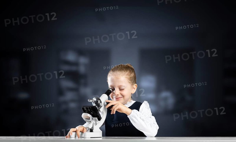 Little girl scientist looking through microscope in classroom at biology lesson. Research and discovery. Formal education in modern school. Schoolgirl in schoolwear making experiment in class