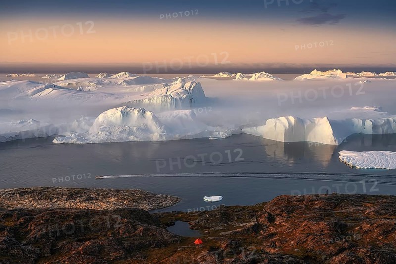 Large glaciers in the bay by the rocky shore in the light of the setting sun with fog over the water with orange tourist tent
