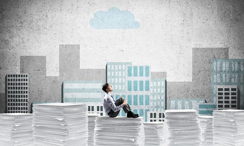 Thoughtful businessman looking away while sitting on pile of documents with drawn cityscape on background. Mixed media