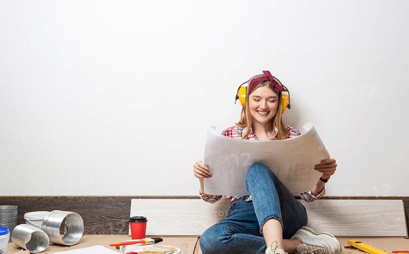 Happy girl in protective headphones sitting on floor with paper blueprint. Home remodeling after moving. Construction tools and materials for building. Young woman wearing checkered shirt and jeans
