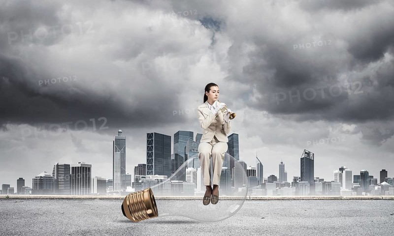 Attractive woman playing trumpet brass on background modern city and storming sky. Young businesslady in white suit sitting on big light bulb with music instrument. Musician practicing and performing