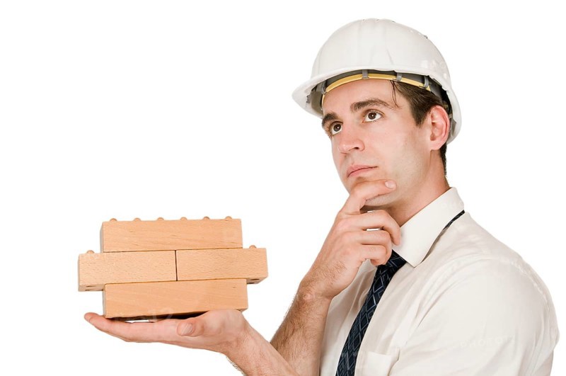 Upper body shot of a young man in a white shirt and white helmet with a stacked wooden block on his left hand and with his right hand on his chin looking upwards in front of a white background