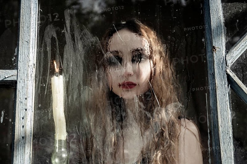 Beautiful sad young girl looking out the window with a candle, good for book cover