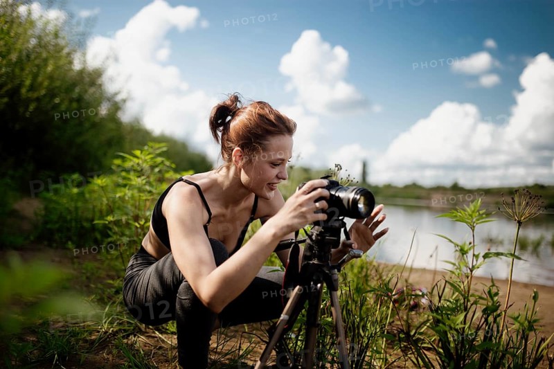 A woman smiles as she adjusts her camera by the lakeside under a blue sky