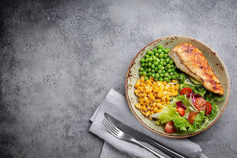 Baked chicken breast on plate with fresh salad, green peas and corn, grey stone background, top view. Healthy fitness meal with chicken fillet, balanced in proteins and carbs. Overhead, copy space