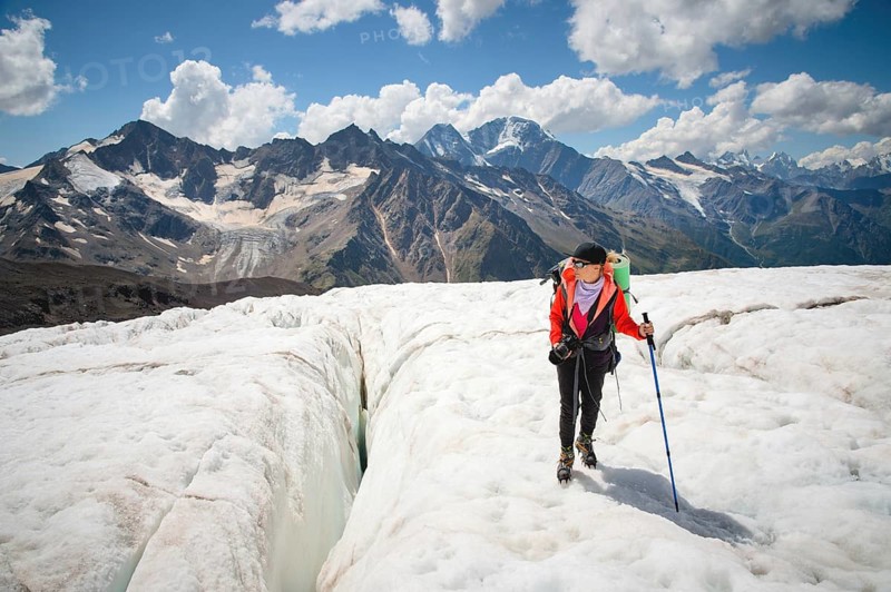 Female mountaineer enjoying the beauty of the glacier walks on the glacier in the crampon and sunglasses. Against the backdrop of the high mountains of the Caucasus sky and clouds